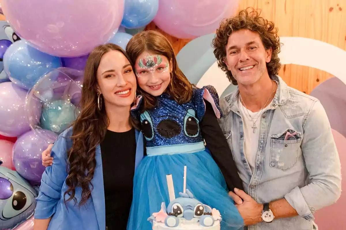 Familia sonriente celebrando una fiesta infantil con temática de Stitch, la niña lleva un vestido azul y maquillaje de fantasía, rodeados de globos y un pastel decorado
