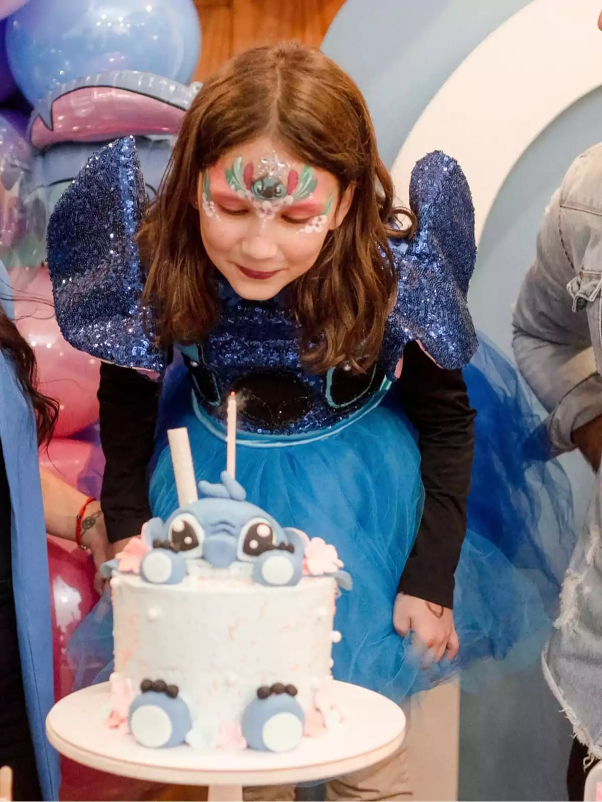 Girl dressed in blue with fantasy makeup blowing out the candles on a cake decorated with a Stitch figure at a children's party Girl dressed in blue with fantasy makeup blowing out the candles on a cake decorated with a Stitch figure at a children's party