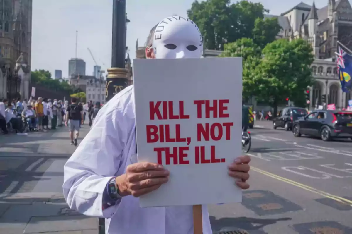 Person wearing a white mask holds a sign that says "Kill the bill not the ill" during a protest on an urban street with buildings and cars in the background.
