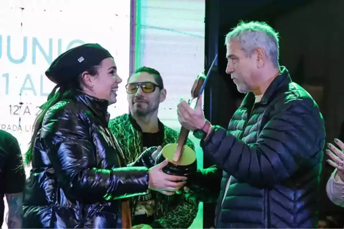 Three people in warm clothing are on a stage while one of them hands an award to another; they all look smiling and there are colorful lights in the atmosphere.