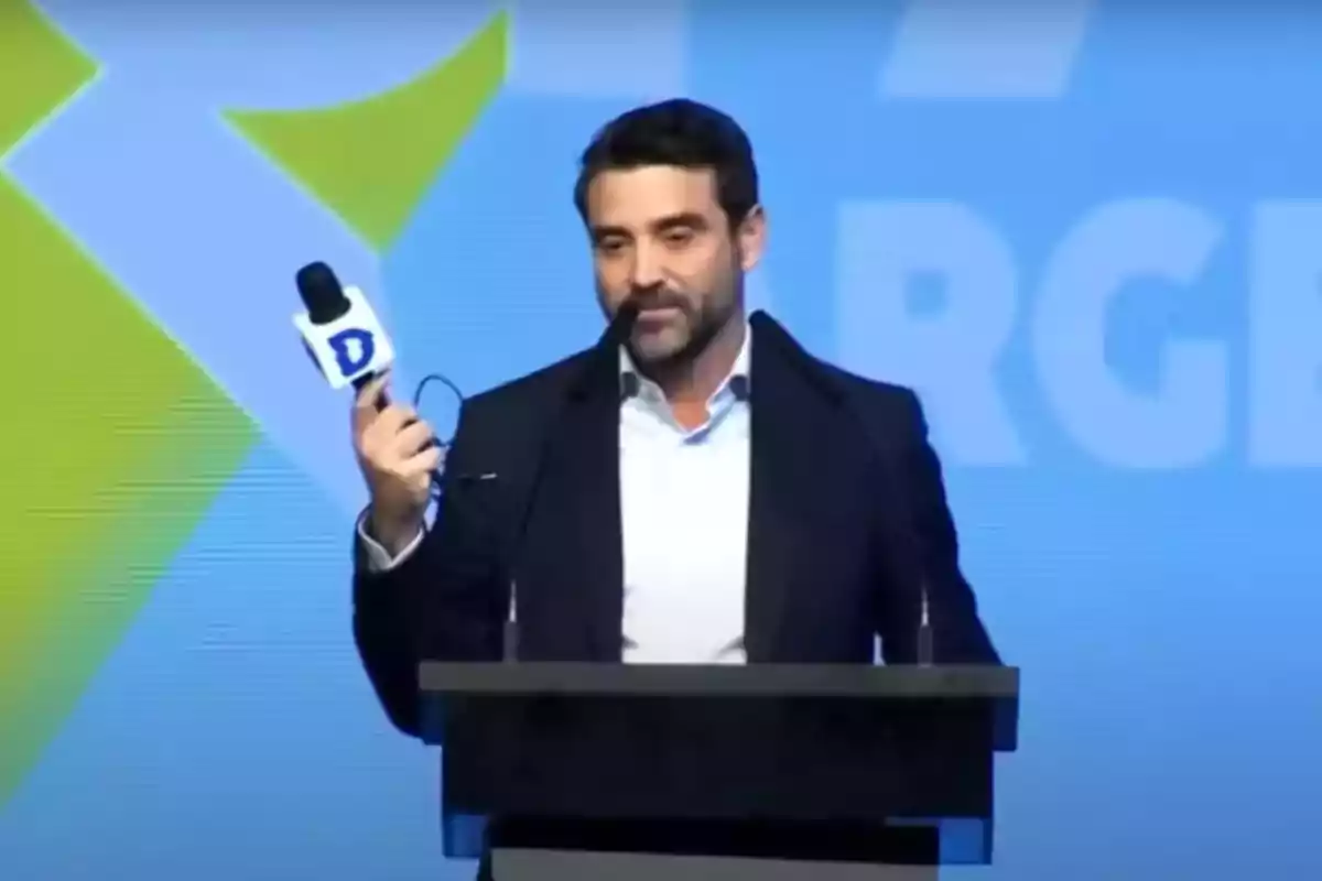 Hombre de barba y cabello oscuro hablando en un podio mientras sostiene un micrófono con una letra D en el fondo de un escenario azul con gráficos geométricos.