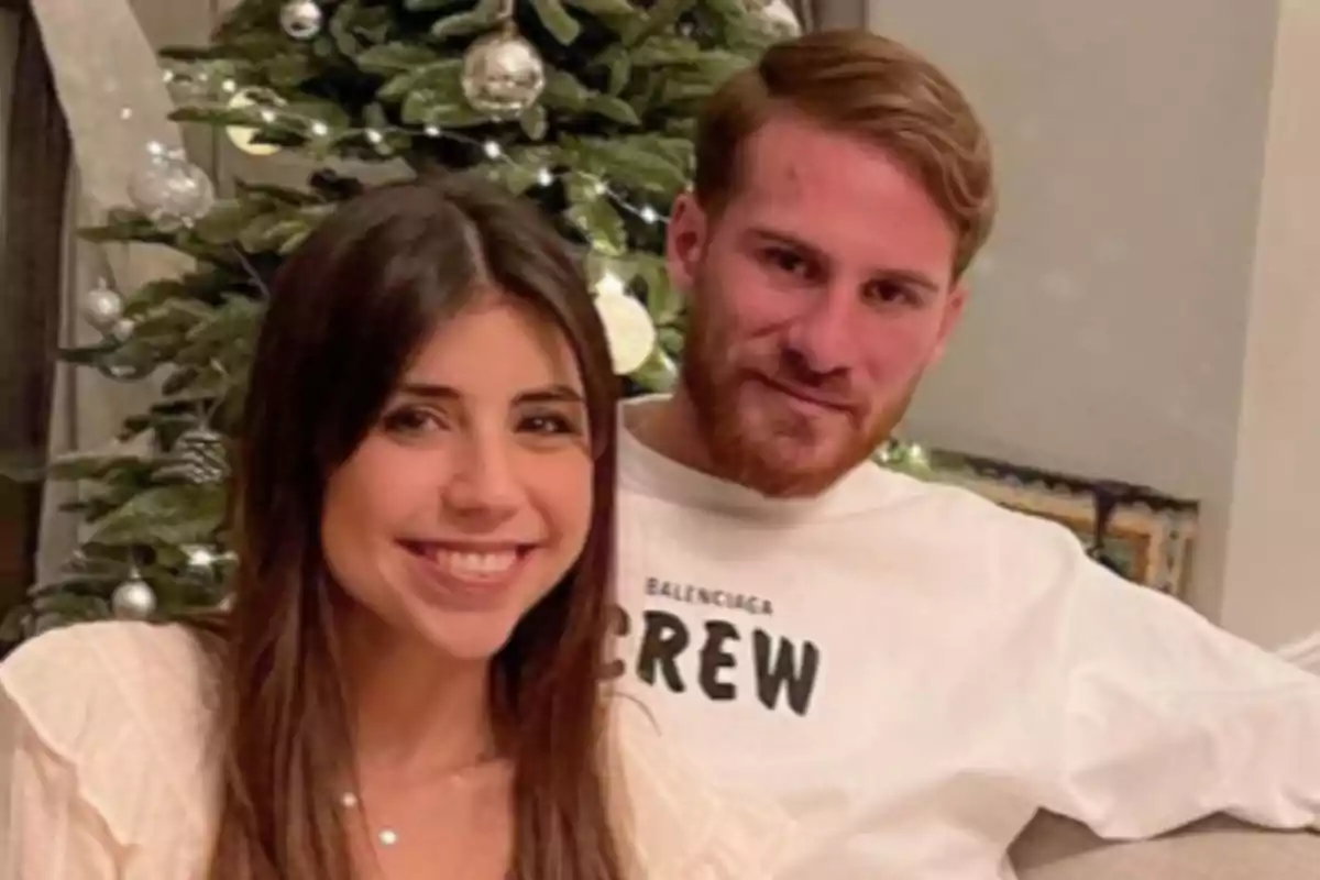 Una pareja sonríe sentada frente a un árbol de Navidad decorado con esferas plateadas Una pareja sonríe sentada frente a un árbol de Navidad decorado con esferas plateadas