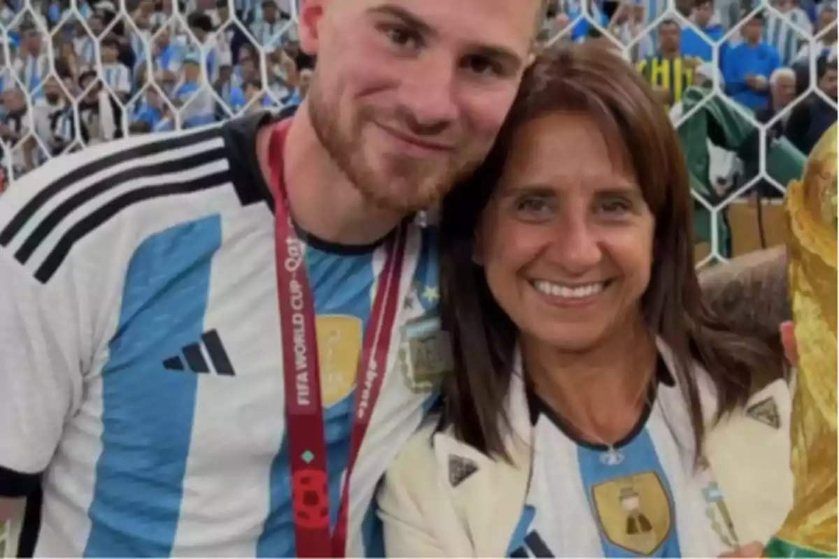 Dos personas sonríen usando camisetas de la selección argentina de fútbol y una de ellas sostiene un trofeo dorado mientras posan frente a una red con público de fondo Dos personas sonríen usando camisetas de la selección argentina de fútbol y una de ellas sostiene un trofeo dorado mientras posan frente a una red con público de fondo
