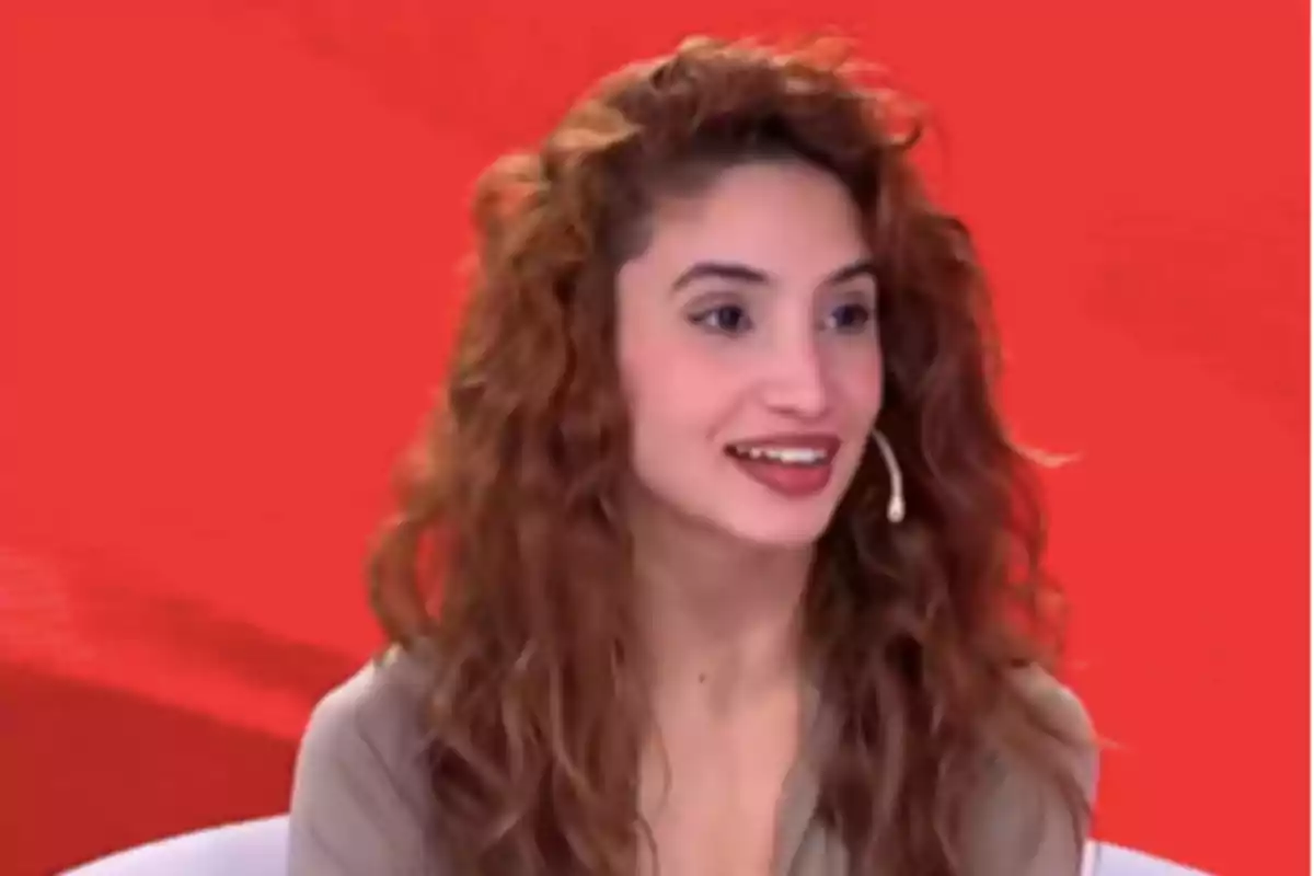 Mujer joven de cabello rizado y largo sentada frente a un fondo rojo usando un micrófono de diadema y sonriendo Mujer joven de cabello rizado y largo sentada frente a un fondo rojo usando un micrófono de diadema y sonriendo