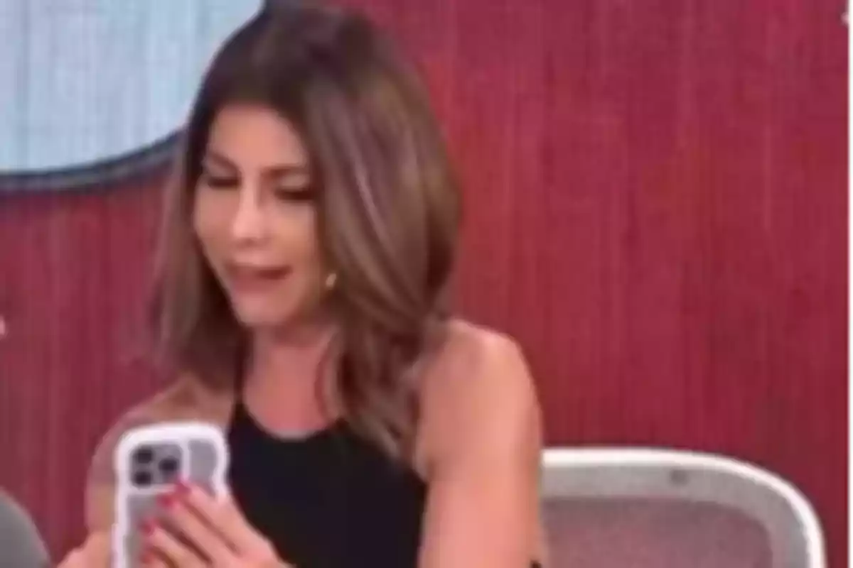 Mujer de cabello castaño y vestido negro mirando un teléfono móvil mientras está sentada frente a un fondo rojo Mujer de cabello castaño y vestido negro mirando un teléfono móvil mientras está sentada frente a un fondo rojo
