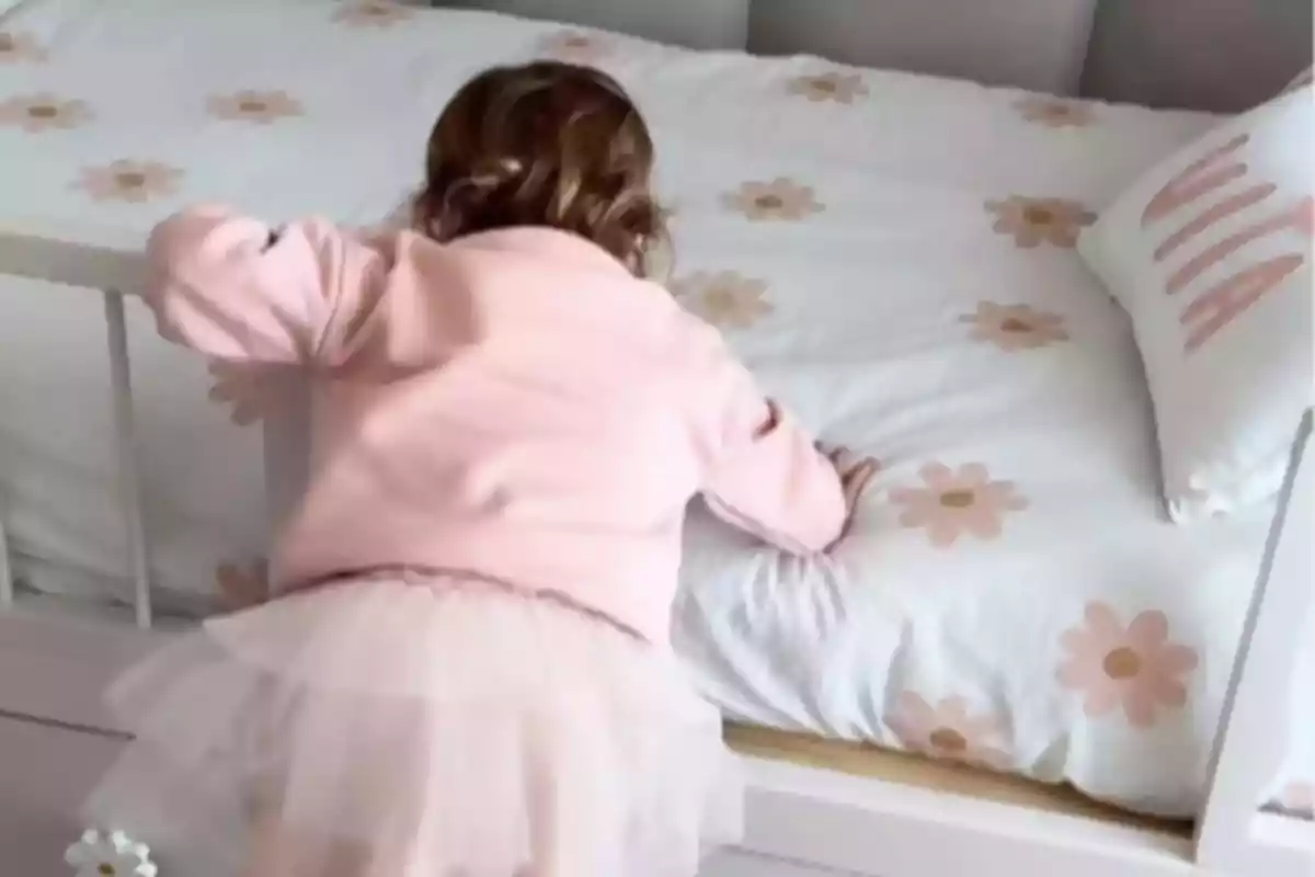 Niña pequeña vestida de rosa subiendo a una cama con sábanas blancas decoradas con flores rosas Niña pequeña vestida de rosa subiendo a una cama con sábanas blancas decoradas con flores rosas
