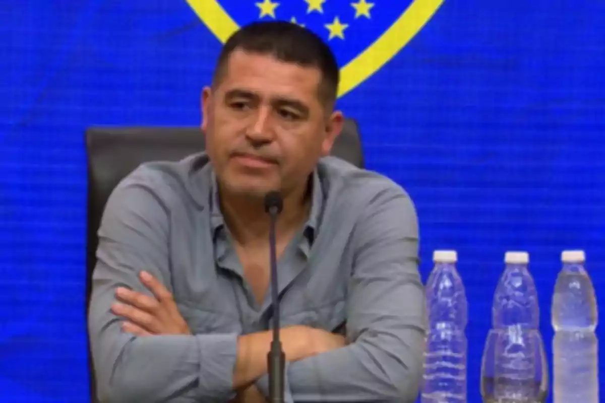 Hombre sentado en una conferencia de prensa con fondo azul y varias botellas de agua sobre la mesa Hombre sentado en una conferencia de prensa con fondo azul y varias botellas de agua sobre la mesa