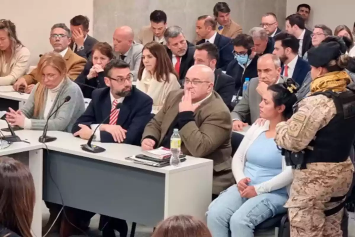 Several people seated in a courtroom, some with microphones in front of them and a woman guarded by a person in military uniform. Several people seated in a courtroom, some with microphones in front of them and a woman guarded by a person in military uniform.