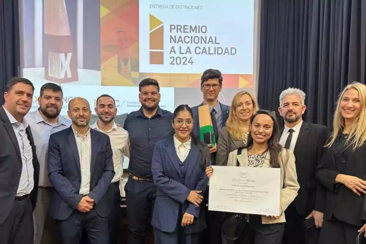 Grupo de personas vestidas de manera formal posando sonrientes en un evento de premiación con un diploma y un trofeo, al fondo se ve una pantalla con el texto Premio Nacional a la Calidad 2024.