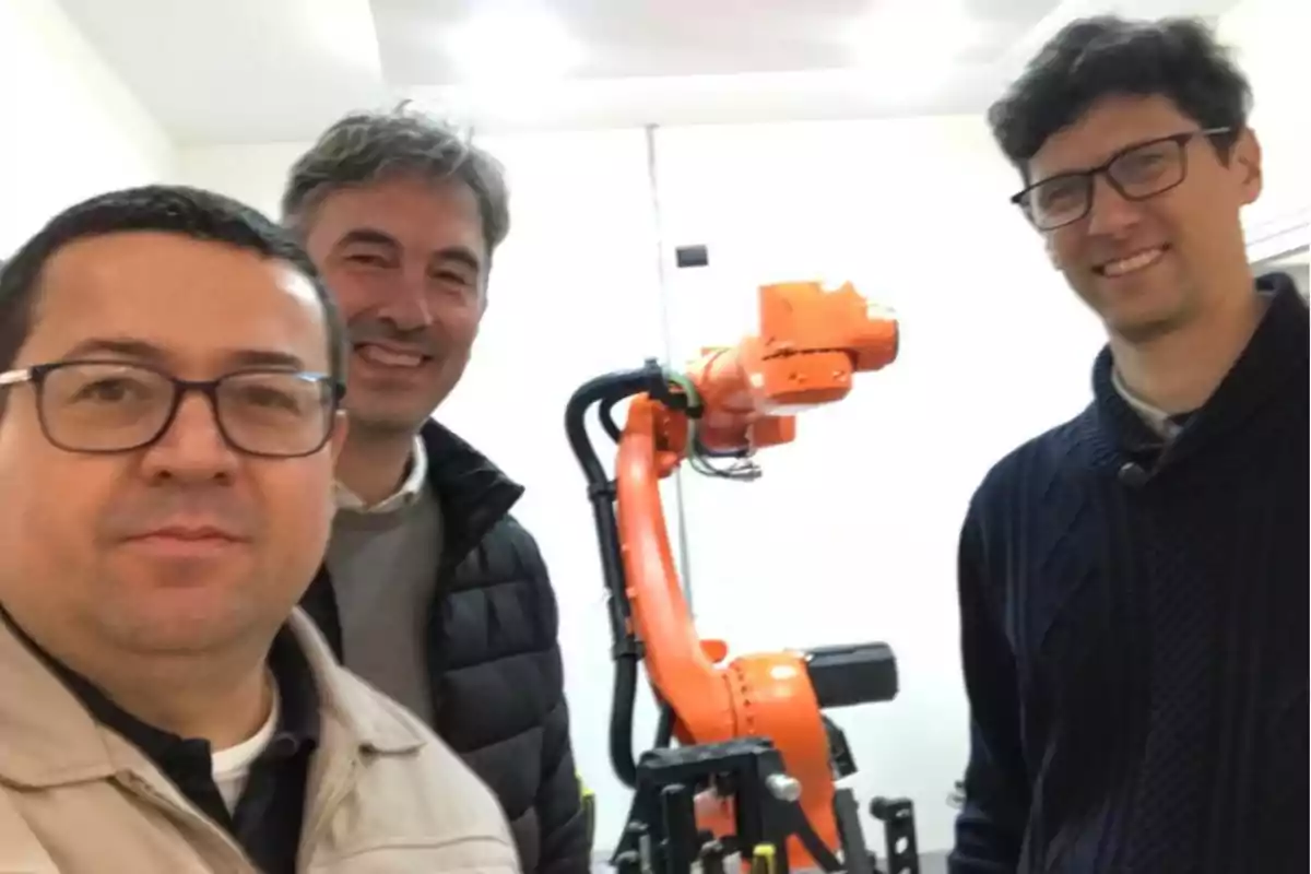 Tres personas posan sonrientes frente a un brazo robótico naranja en un entorno interior iluminado Tres personas posan sonrientes frente a un brazo robótico naranja en un entorno interior iluminado