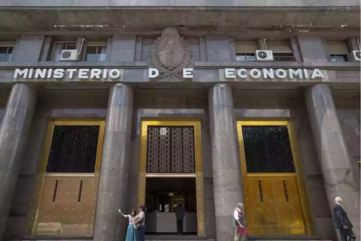 Fachada del edificio del Ministerio de Economía de Argentina con columnas de mármol y puertas doradas, personas caminando y tomándose una selfie en la entrada. Fachada del edificio del Ministerio de Economía de Argentina con columnas de mármol y puertas doradas, personas caminando y tomándose una selfie en la entrada.