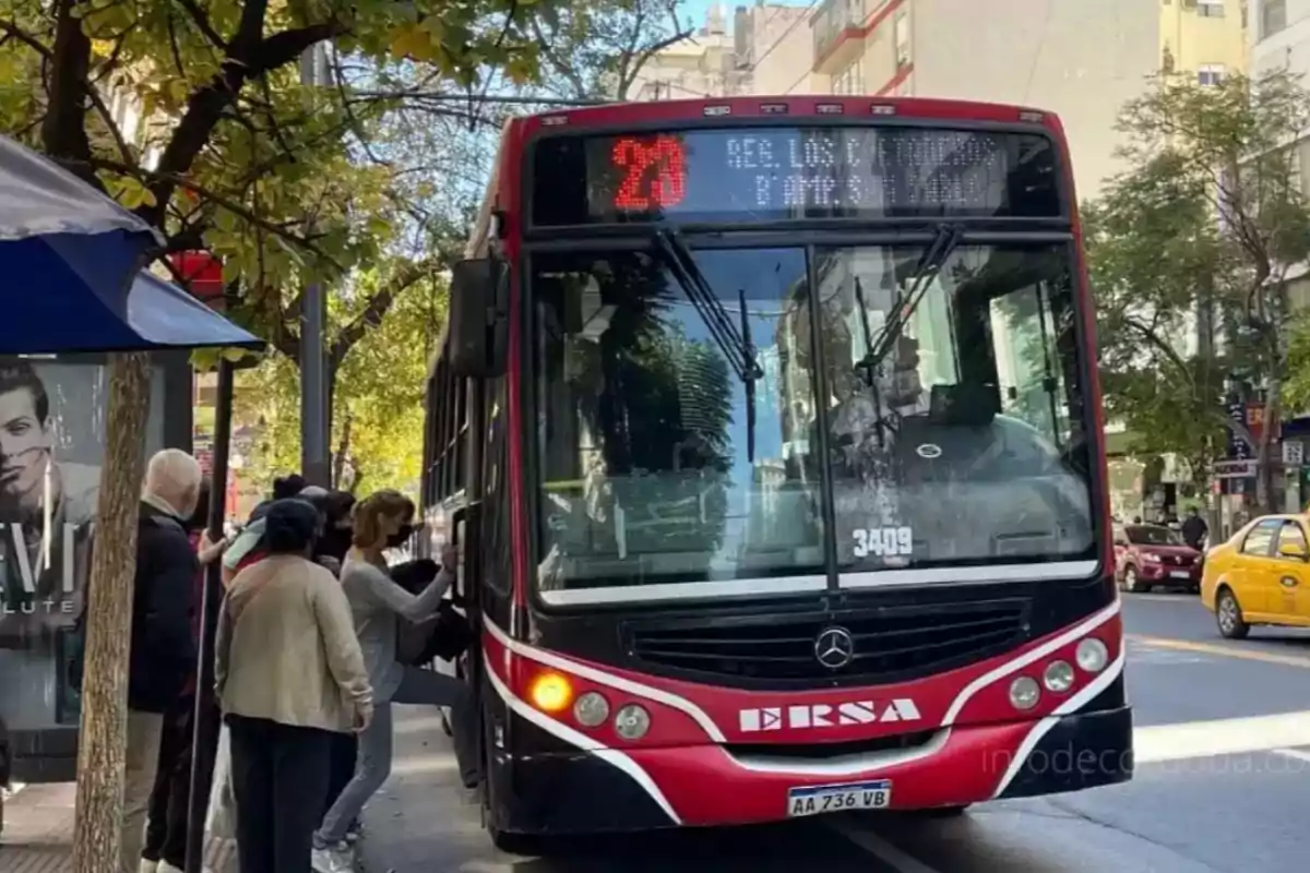 A red and black bus stopped at a bus stop while several people get on in an urban street with trees and buildings around.