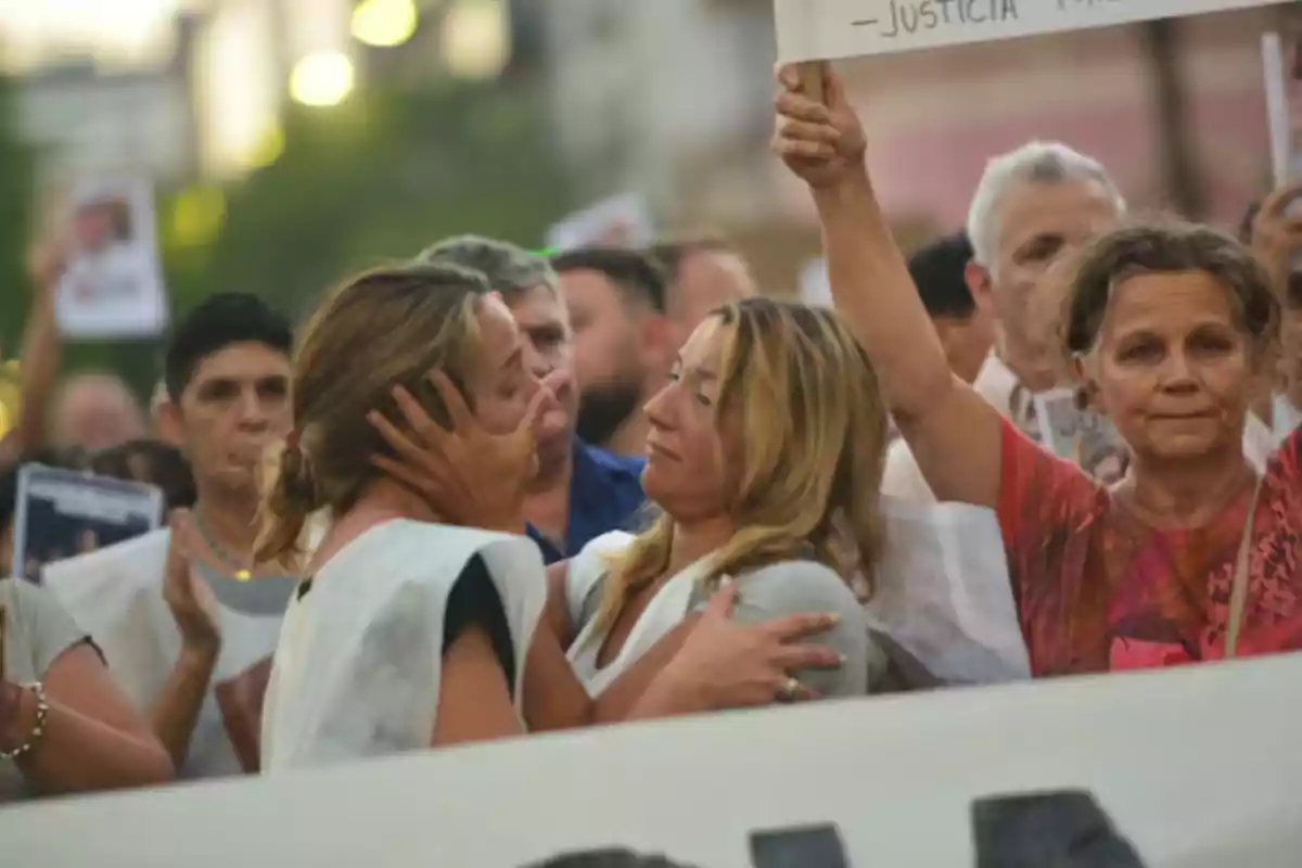 Un grupo de personas participa en una manifestación, dos mujeres se abrazan emocionadas mientras otra sostiene un cartel y el ambiente transmite sentimientos de apoyo y solidaridad. Un grupo de personas participa en una manifestación, dos mujeres se abrazan emocionadas mientras otra sostiene un cartel y el ambiente transmite sentimientos de apoyo y solidaridad.