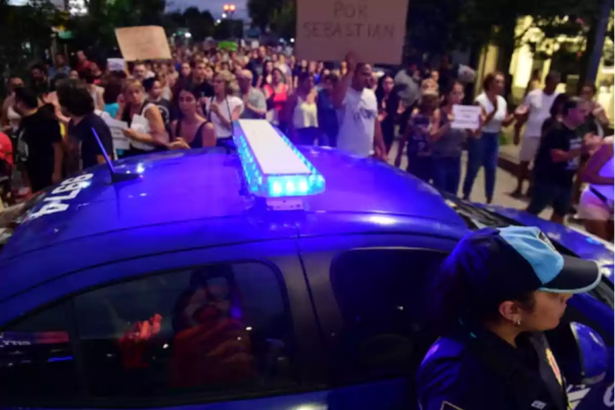 Una multitud de personas marcha por la calle detrás de un auto de policía con luces encendidas mientras una oficial observa la manifestación. Una multitud de personas marcha por la calle detrás de un auto de policía con luces encendidas mientras una oficial observa la manifestación.