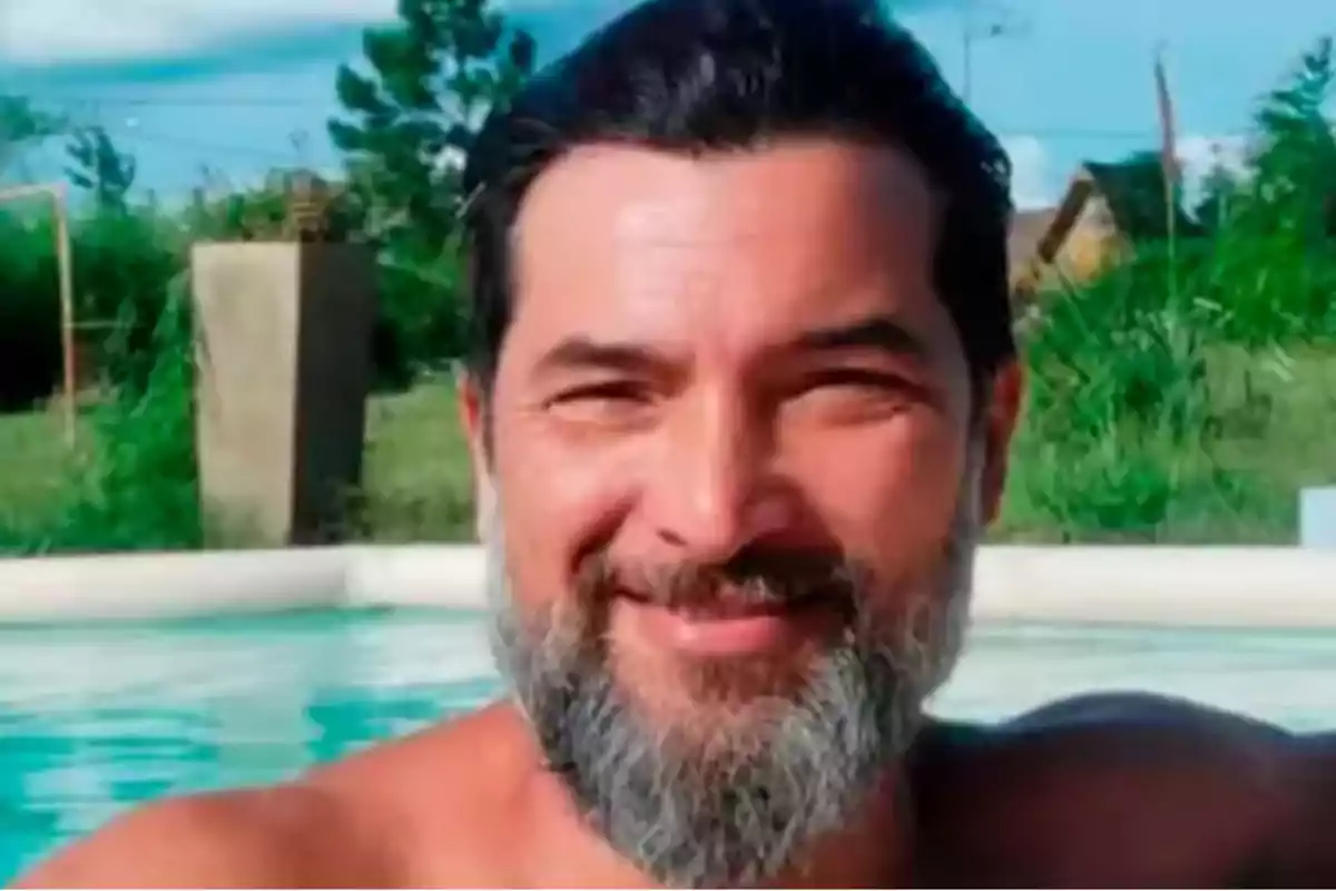 Hombre de barba y cabello oscuro sonriendo dentro de una piscina al aire libre con vegetación de fondo Hombre de barba y cabello oscuro sonriendo dentro de una piscina al aire libre con vegetación de fondo