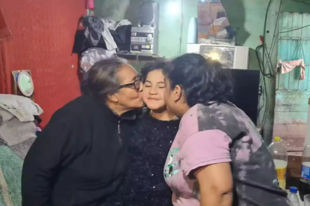 Three people in a room, a girl in the center smiles with her eyes closed while an older woman and another woman kiss her on the cheeks.