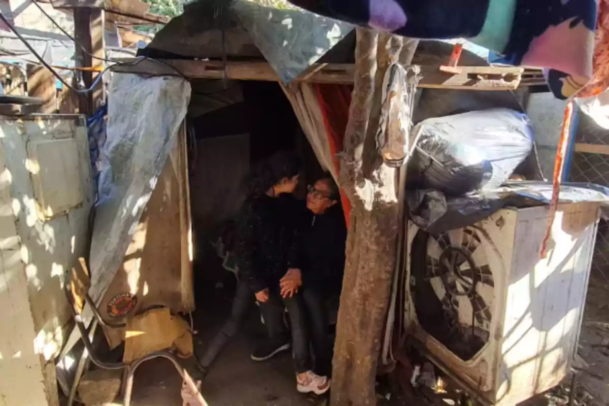 A girl and an older woman look at each other and hold hands at the entrance of an improvised dwelling made of tarps and wood, surrounded by old objects and a tree. A girl and an older woman look at each other and hold hands at the entrance of an improvised dwelling made of tarps and wood, surrounded by old objects and a tree.