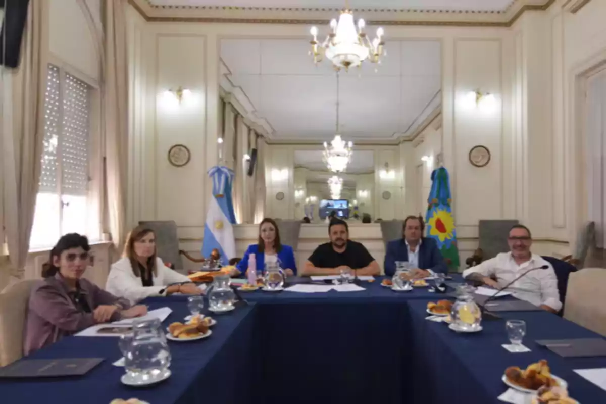Siete personas sentadas alrededor de una mesa en una sala elegante con banderas y lámparas de araña, participando en una reunión formal con documentos y comida sobre la mesa. Siete personas sentadas alrededor de una mesa en una sala elegante con banderas y lámparas de araña, participando en una reunión formal con documentos y comida sobre la mesa.
