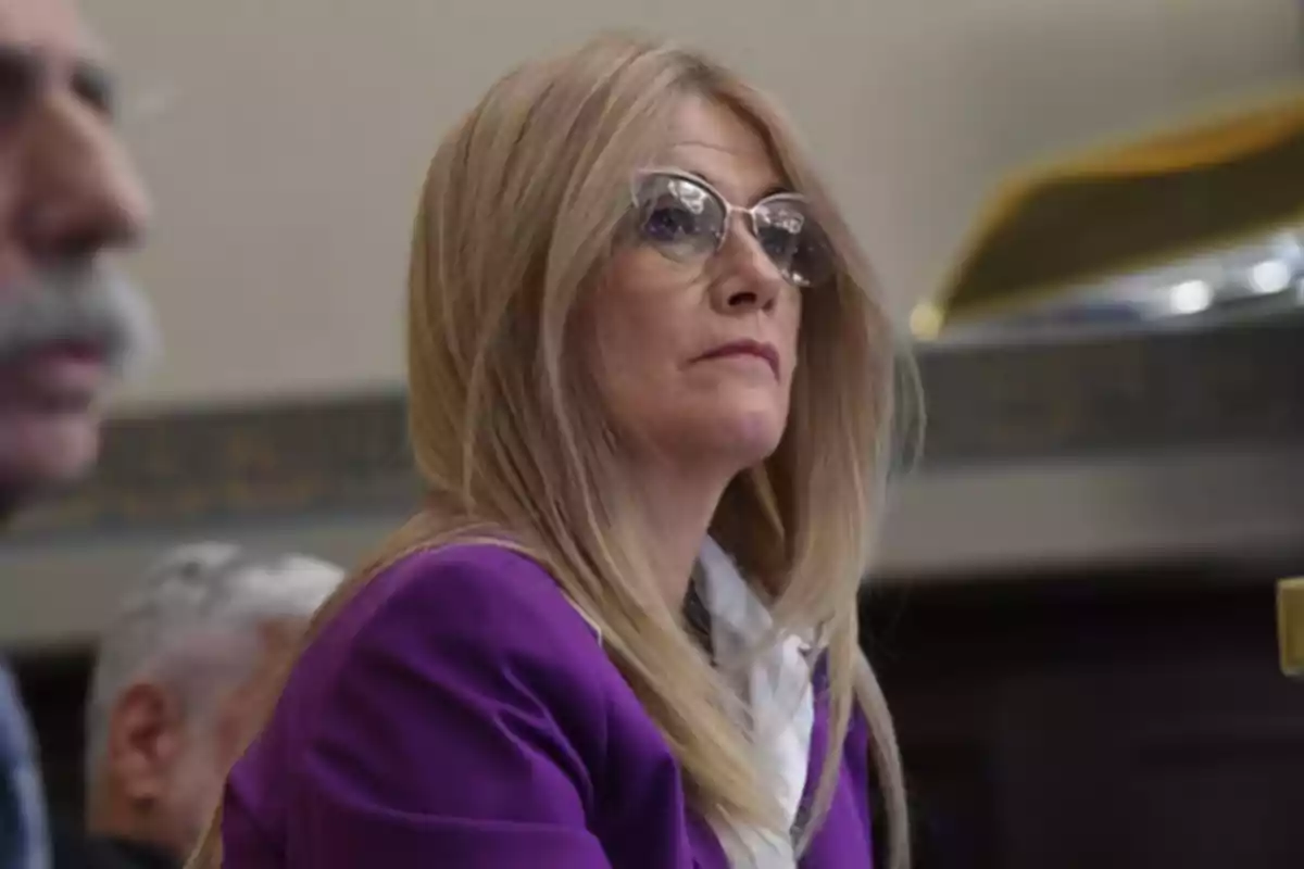 Mujer de cabello rubio y lacio con gafas y saco morado sentada en un lugar cerrado Mujer de cabello rubio y lacio con gafas y saco morado sentada en un lugar cerrado