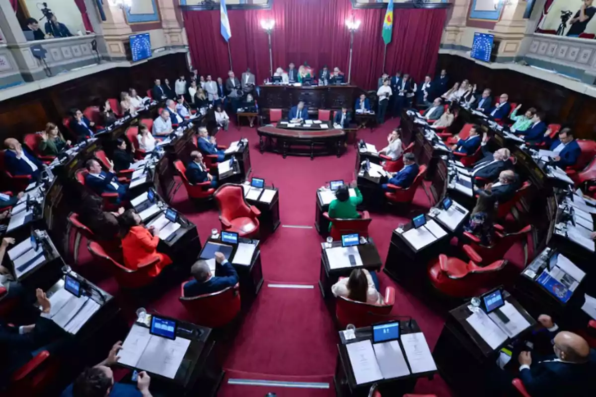 Vista general de una sesión parlamentaria con legisladores sentados en sus bancas dentro de un recinto con alfombra roja y cortinas, mientras algunas personas observan desde los palcos superiores