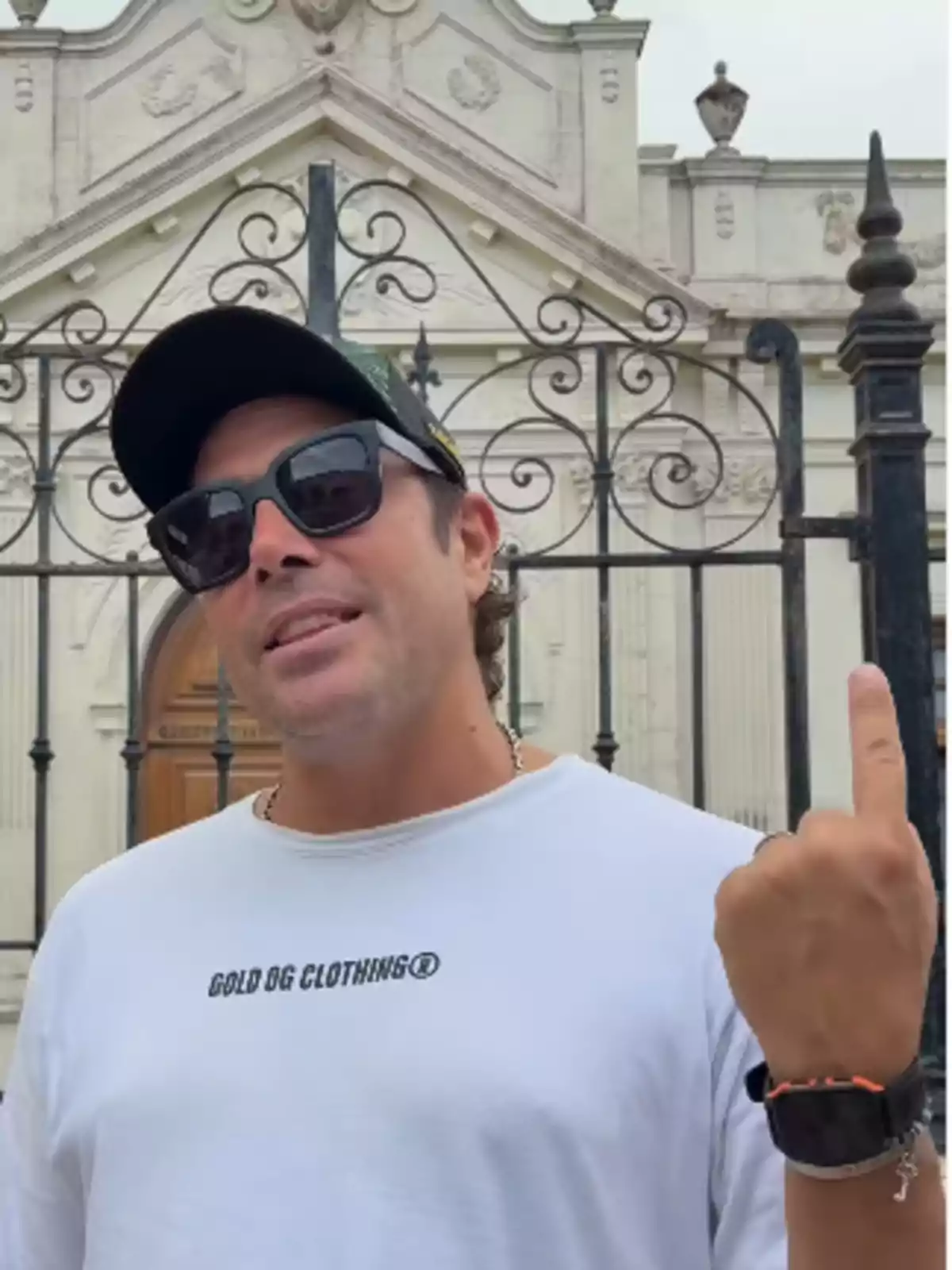 Hombre con gafas de sol y gorra blanca posando frente a una reja de hierro y un edificio antiguo mientras señala hacia arriba con el dedo índice Hombre con gafas de sol y gorra blanca posando frente a una reja de hierro y un edificio antiguo mientras señala hacia arriba con el dedo índice