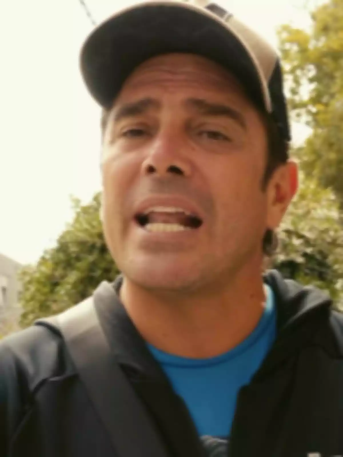 Hombre con gorra y chaqueta negra hablando al aire libre con árboles de fondo Hombre con gorra y chaqueta negra hablando al aire libre con árboles de fondo