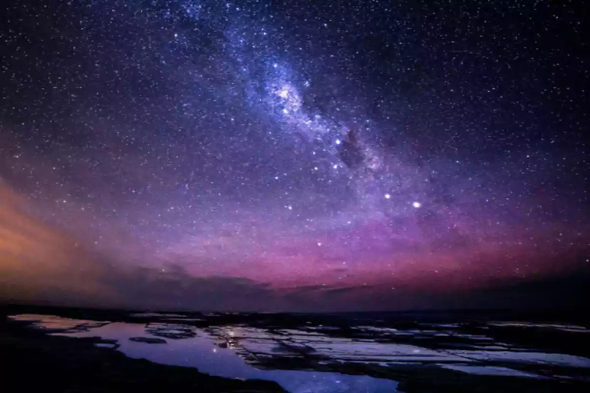 Cielo nocturno estrellado con la Vía Láctea visible sobre un paisaje con agua reflejando las estrellas