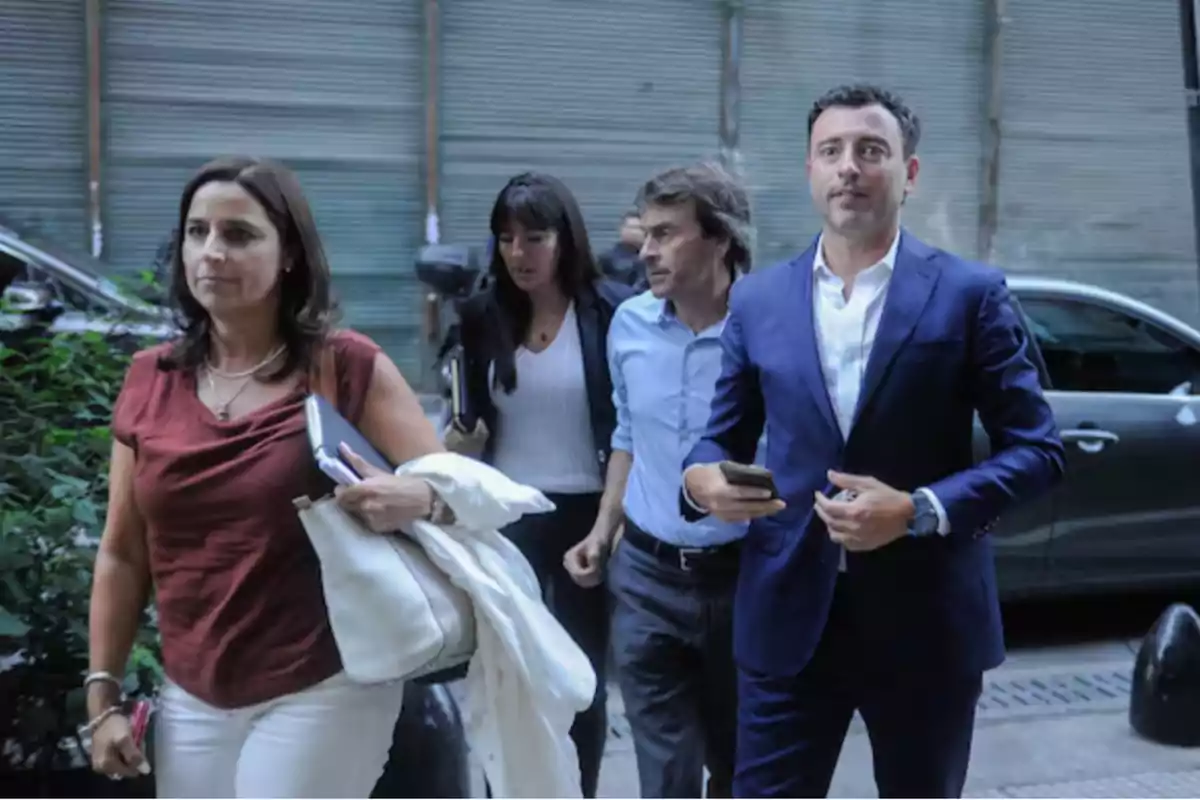 Cuatro personas caminando por la calle junto a un auto y un portón metálico al fondo Cuatro personas caminando por la calle junto a un auto y un portón metálico al fondo