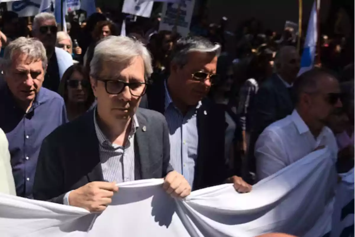 Un grupo de personas marcha en la calle sosteniendo una pancarta blanca mientras otras personas los acompañan detrás Un grupo de personas marcha en la calle sosteniendo una pancarta blanca mientras otras personas los acompañan detrás