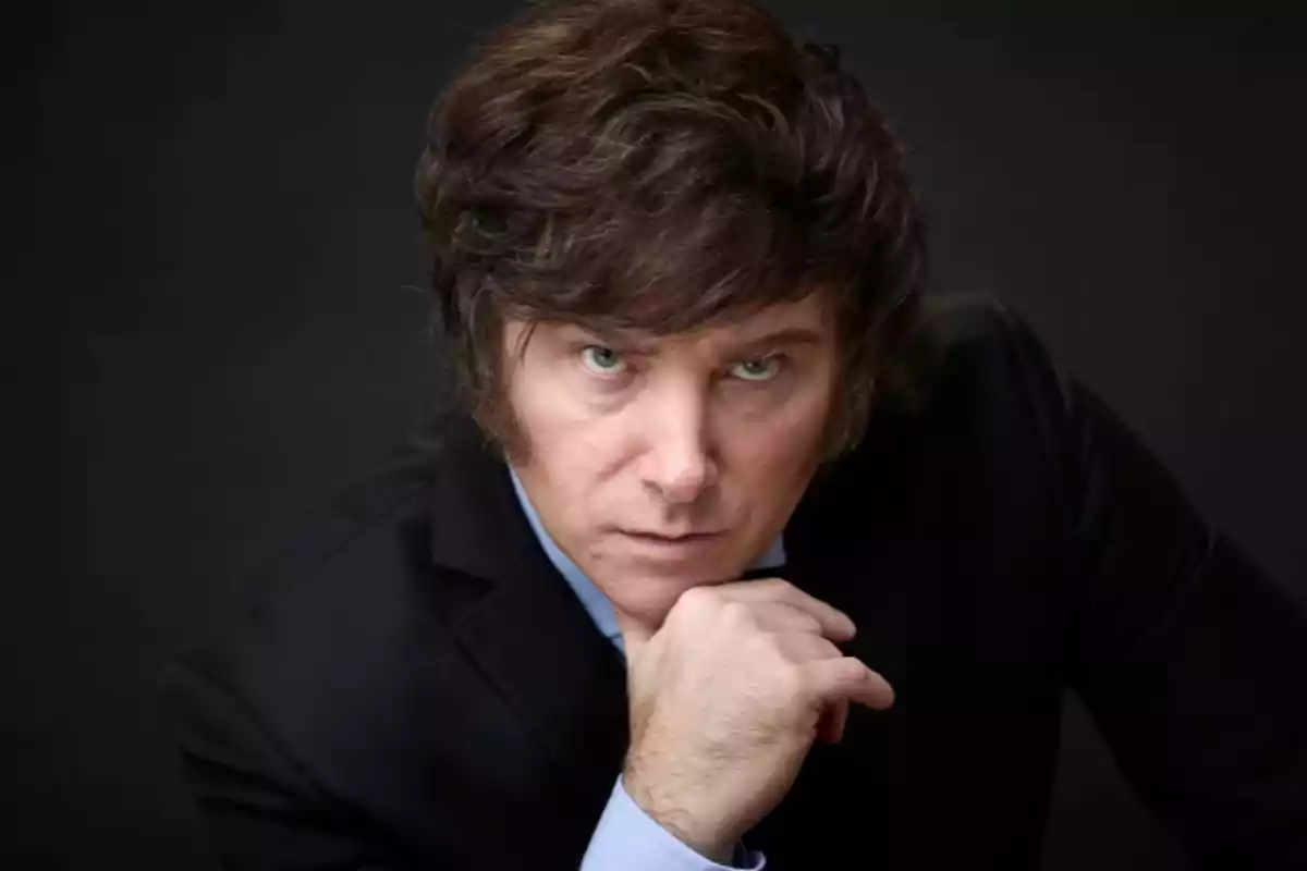 Hombre de cabello castaño y ojos claros con expresión seria apoyando su barbilla en la mano y vistiendo saco negro sobre fondo oscuro Hombre de cabello castaño y ojos claros con expresión seria apoyando su barbilla en la mano y vistiendo saco negro sobre fondo oscuro
