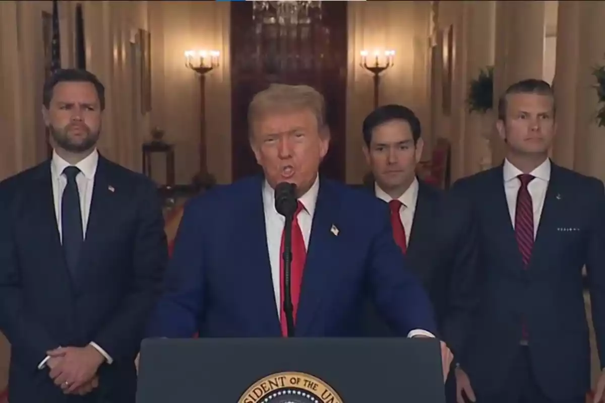 A man in a blue suit and red tie speaks in front of a microphone with three men in suits behind him in an elegant room with lit chandeliers in the background.