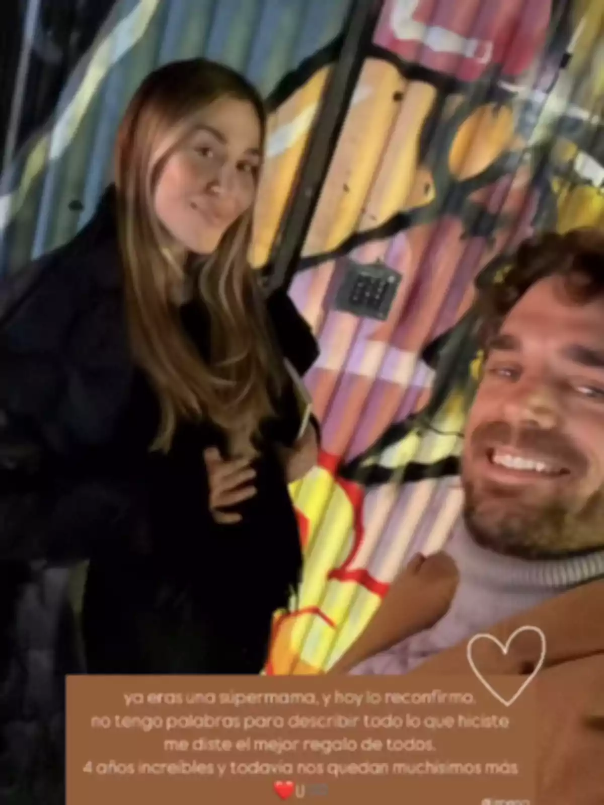 Una pareja sonríe frente a una pared con arte urbano colorido, mientras la mujer posa con la mano sobre su vientre y ambos parecen felices. Una pareja sonríe frente a una pared con arte urbano colorido, mientras la mujer posa con la mano sobre su vientre y ambos parecen felices.