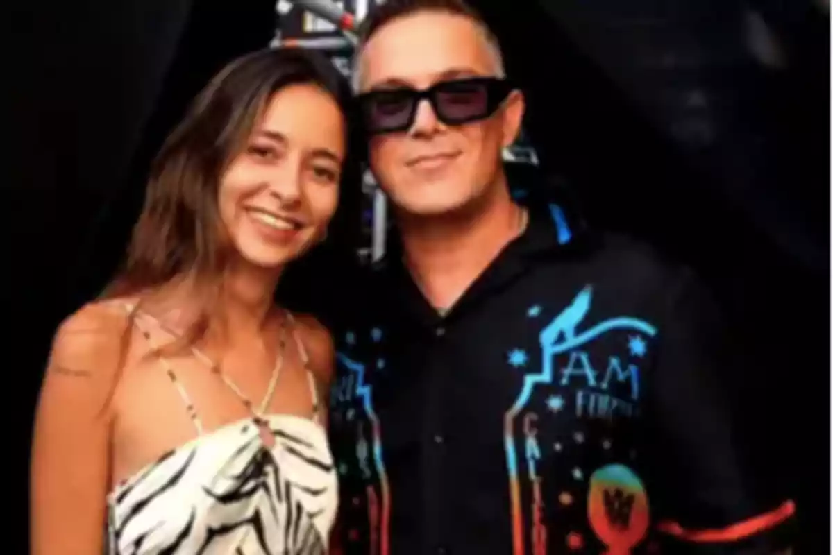 Una mujer joven con vestido blanco de estampado negro sonríe junto a un hombre con gafas oscuras y camisa negra con detalles de colores, ambos posan frente a un fondo oscuro. Una mujer joven con vestido blanco de estampado negro sonríe junto a un hombre con gafas oscuras y camisa negra con detalles de colores, ambos posan frente a un fondo oscuro.