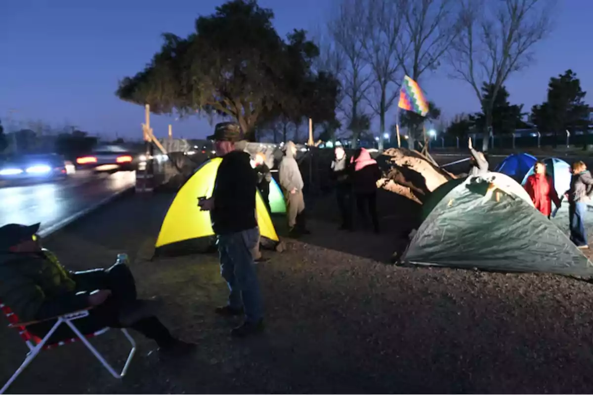 People camping by a roadside at night with several tents and a colorful flag waving, while some cars pass by in the background. People camping by a roadside at night with several tents and a colorful flag waving, while some cars pass by in the background.