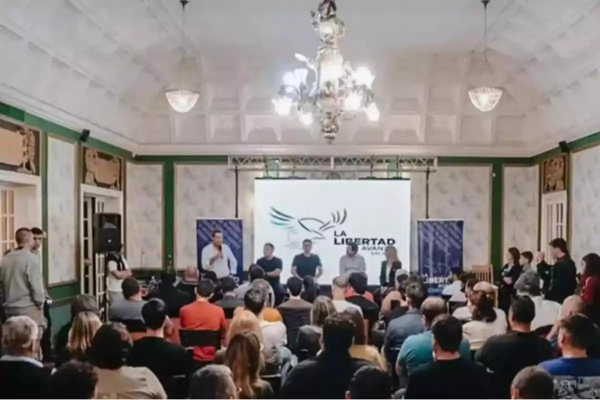 A group of people attends a conference in an elegant hall with chandeliers and decorated walls; in the background, there is a screen with the La Libertad Avanza logo.