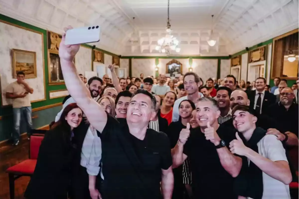 A large group of people smiles while taking a selfie in an elegant room with lamps and paintings on the walls.