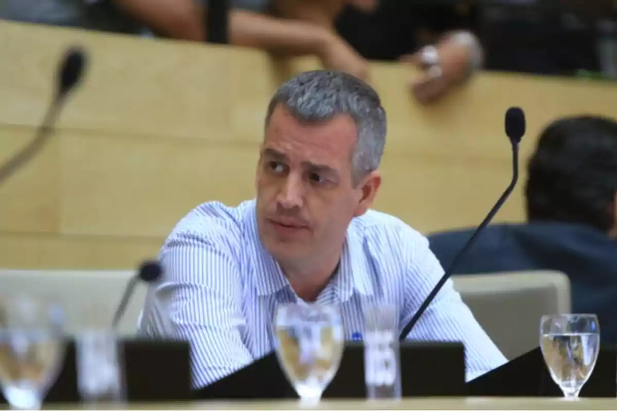 Short-haired, gray-haired man sitting in a conference room with a microphone and water glasses on the table Short-haired, gray-haired man sitting in a conference room with a microphone and water glasses on the table
