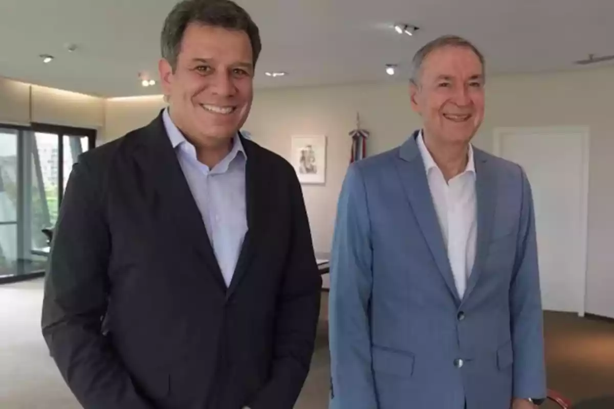 Two men standing in an office, both smiling and wearing jackets, one dark-colored and the other light blue.