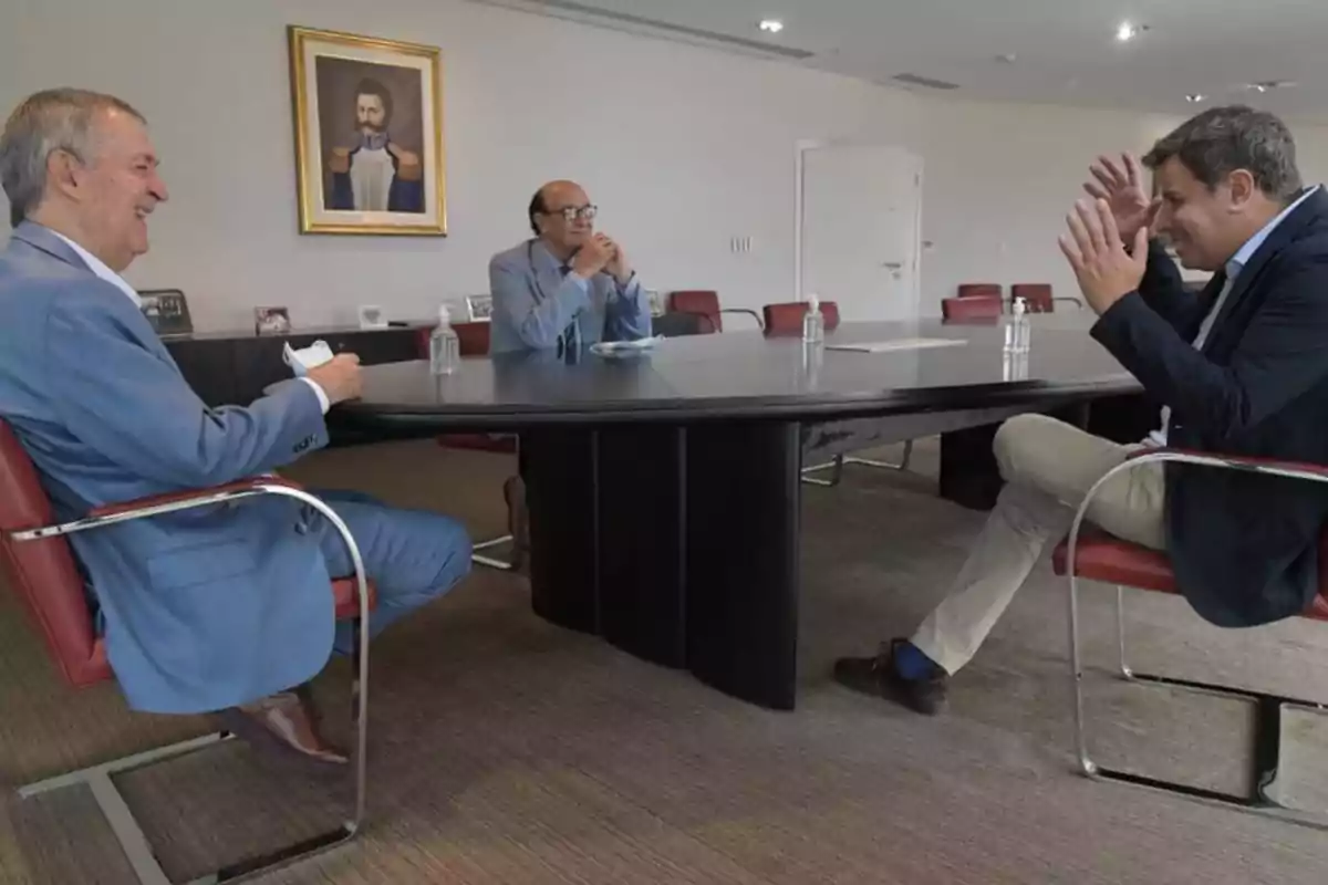 Three men are having an animated conversation while sitting around a table in a modern office, with a portrait on the wall in the background. Three men are having an animated conversation while sitting around a table in a modern office, with a portrait on the wall in the background.