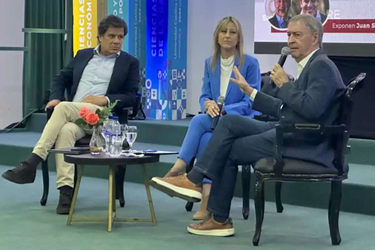 Three people are seated on a stage participating in a talk; one of them is holding a microphone while the other two listen alertly. There is a small table with glasses and a bottle of water in front of them. Three people are seated on a stage participating in a talk; one of them is holding a microphone while the other two listen alertly. There is a small table with glasses and a bottle of water in front of them.