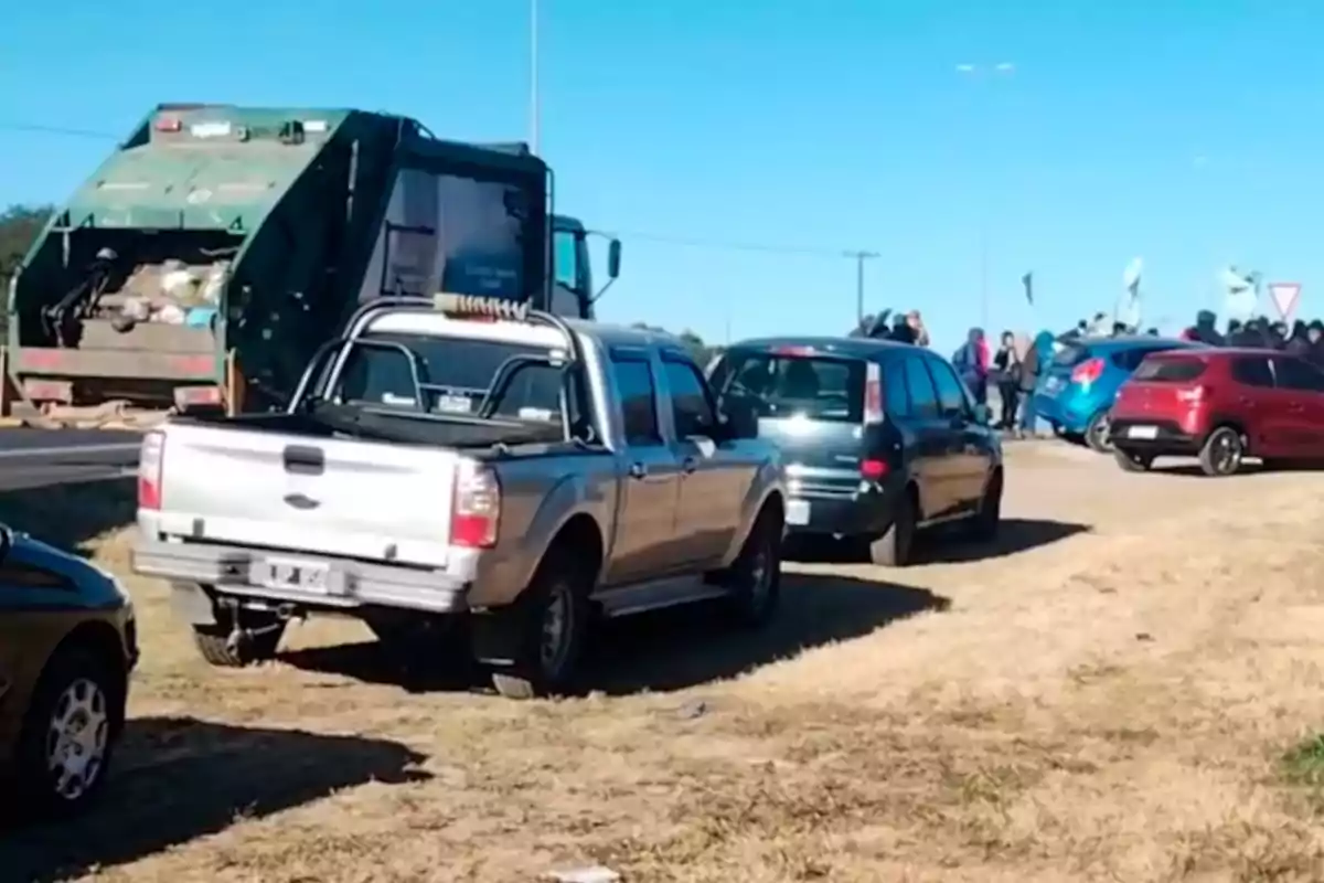 Varios autos y una camioneta están detenidos junto a un camión de basura en una calle de tierra mientras un grupo de personas se reúne al fondo bajo un cielo despejado.