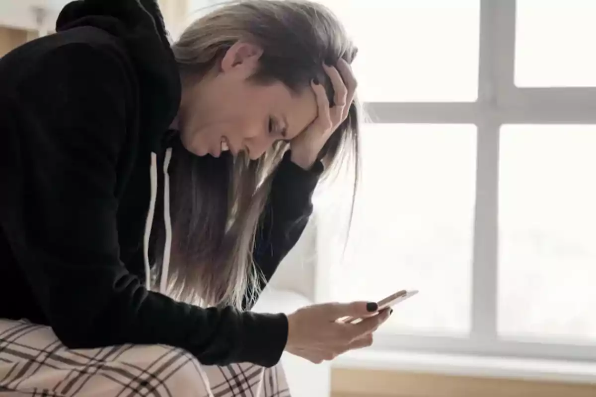 Woman sitting with a distressed expression, looking at her cell phone and holding her head with one hand Woman sitting with a distressed expression, looking at her cell phone and holding her head with one hand