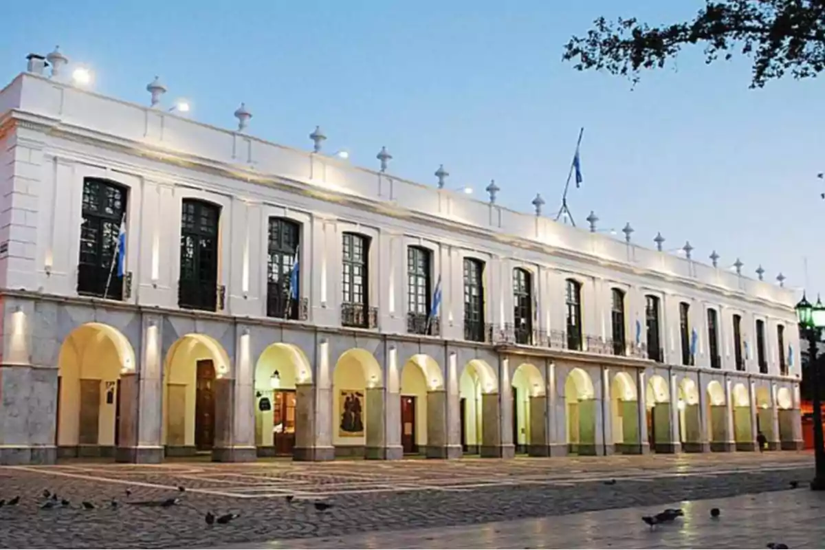 Two-story historic building with arches on the ground floor and large windows above, illuminated at sunset in a plaza with some pigeons on the ground Two-story historic building with arches on the ground floor and large windows above, illuminated at sunset in a plaza with some pigeons on the ground