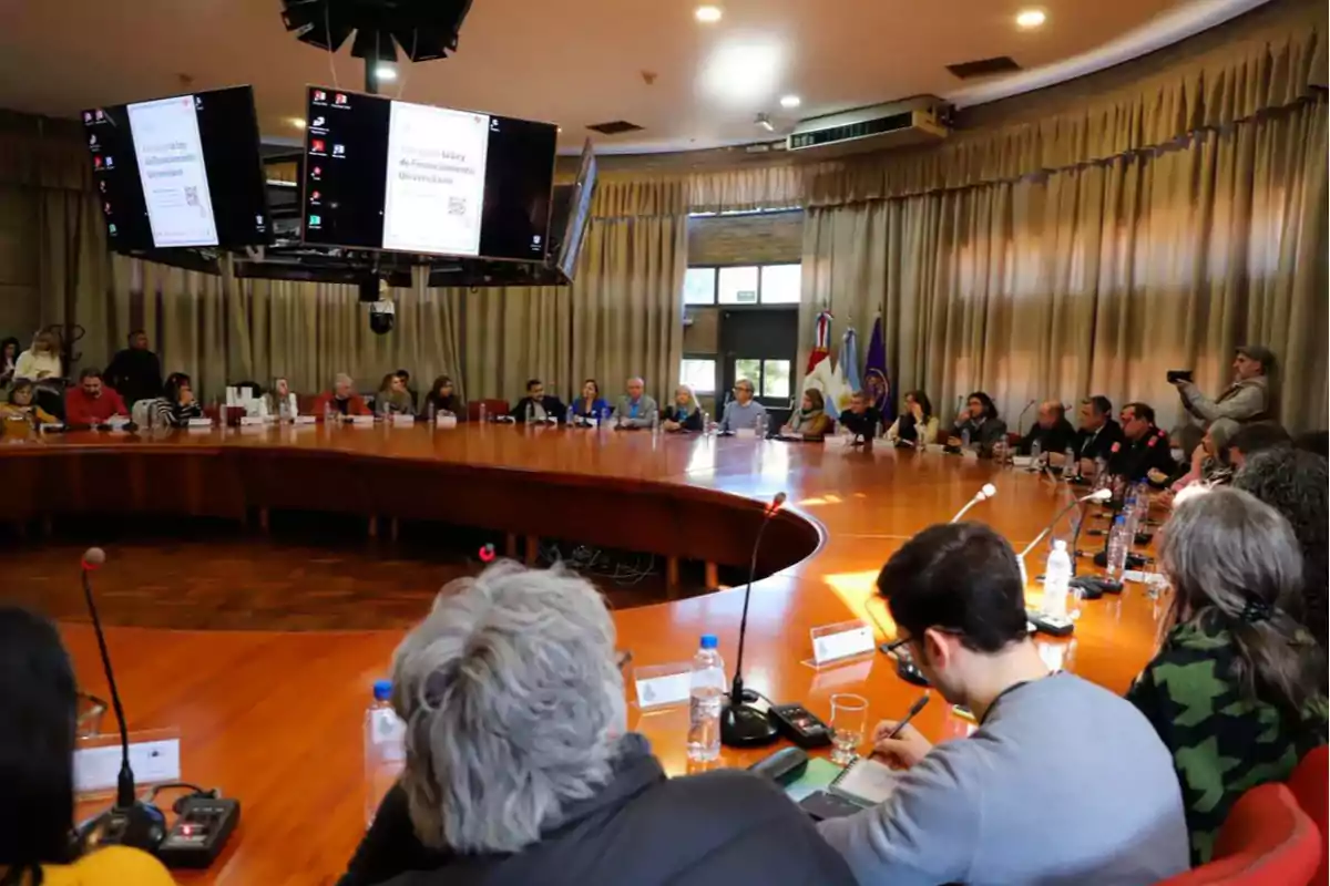 People gathered around a large oval table in a conference room with screens and microphones