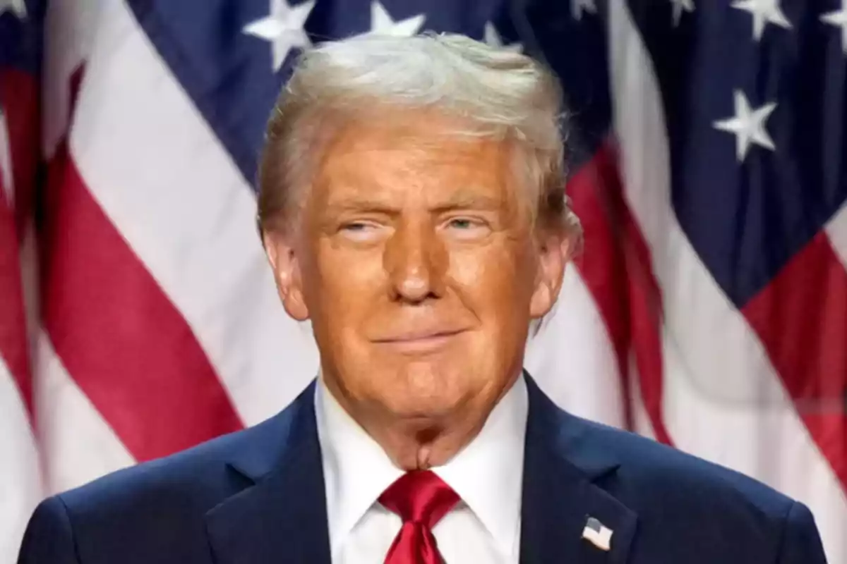 Hombre de cabello rubio con traje oscuro y corbata roja frente a banderas de Estados Unidos
