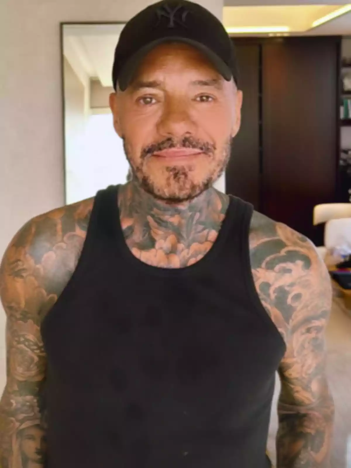 Hombre con barba y tatuajes vistiendo una camiseta negra sin mangas y gorra negra en una habitación moderna Hombre con barba y tatuajes vistiendo una camiseta negra sin mangas y gorra negra en una habitación moderna