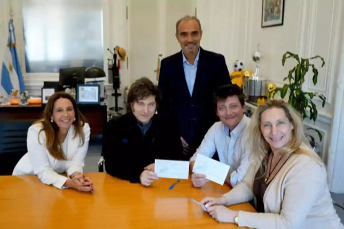 Cinco personas sonríen alrededor de una mesa en una oficina con banderas y trofeos al fondo dos de ellas sostienen papeles en la mano Cinco personas sonríen alrededor de una mesa en una oficina con banderas y trofeos al fondo dos de ellas sostienen papeles en la mano