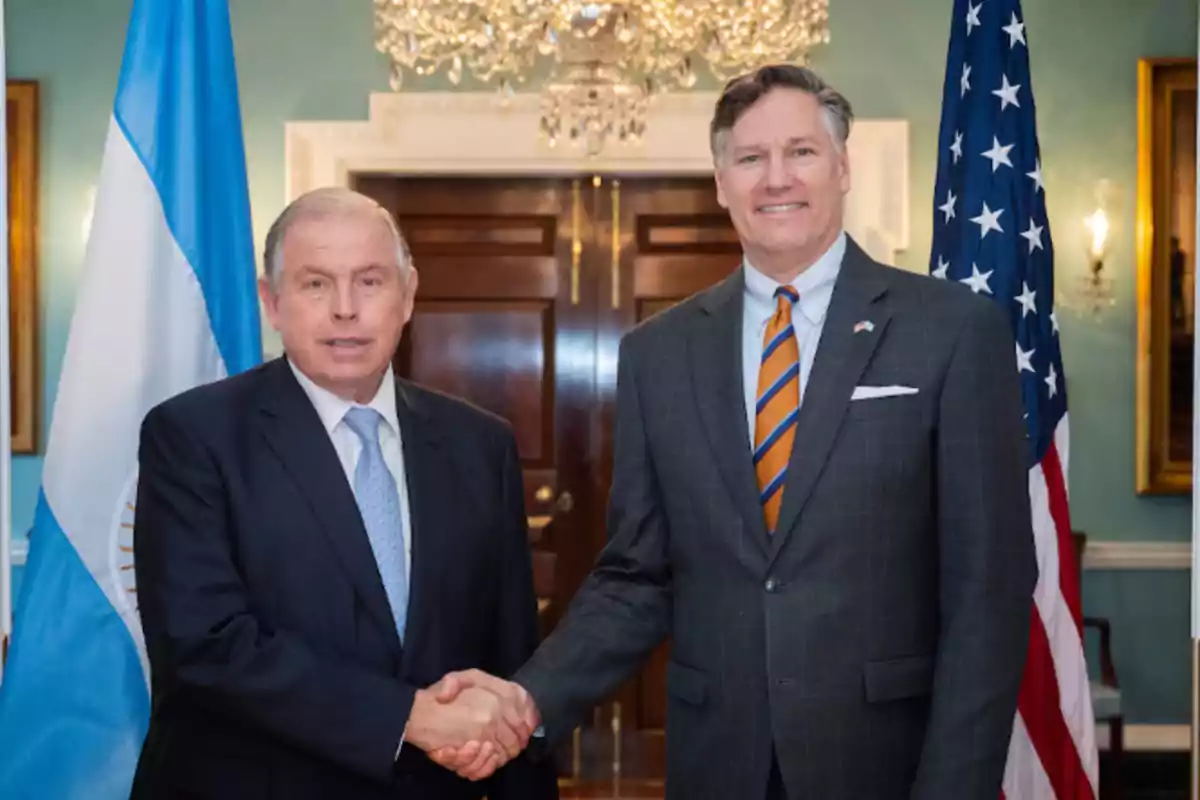 Two men in suits shake hands in front of the flags of Argentina and the United States in a formal setting.