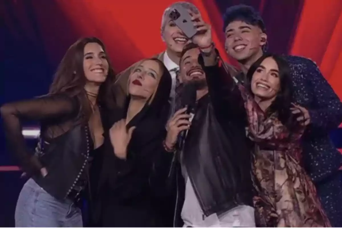 Grupo de seis personas sonríe mientras se toman una selfie en un escenario iluminado con luces rojas de fondo Grupo de seis personas sonríe mientras se toman una selfie en un escenario iluminado con luces rojas de fondo