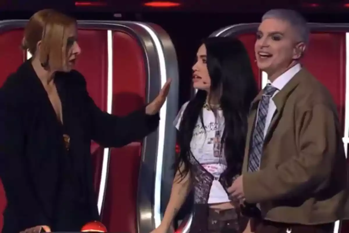 Tres personas interactúan en un set de televisión con sillas rojas al fondo, una de ellas parece estar hablando de manera seria mientras las otras dos observan con expresiones de sorpresa y sonrisa. Tres personas interactúan en un set de televisión con sillas rojas al fondo, una de ellas parece estar hablando de manera seria mientras las otras dos observan con expresiones de sorpresa y sonrisa.